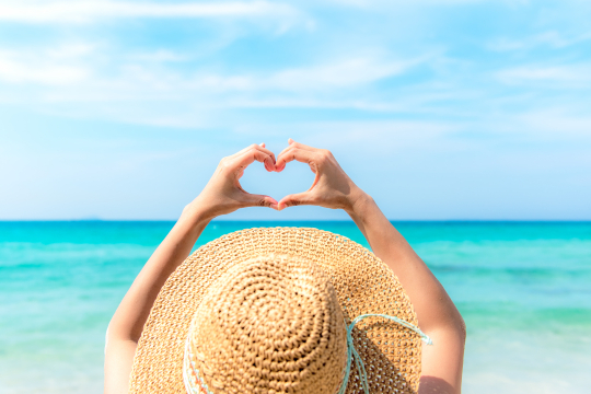 Frau mit gro&szlig;em Sonnenhut von hinten streckt ihre H&auml;nde nach oben und macht eine Herzform damit, im Hintergrund das Meer
